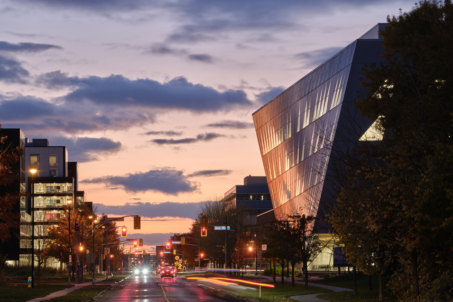 Street view at dusk showing modern, angular buildings with large glass facades, illuminated by the warm glow of interior lights, under a colorful evening sky.